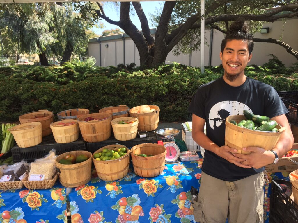 joseph from mandela partners standing at an organic produce stand holding a basket of cucumbers